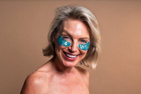 Beauty portrait of beautiful mature woman, concepts about cosmetics, skincare and wellness. Happy mature woman with blond hair and bare shoulders smiling while wearing blue hydrogel eye patchesの写真素材