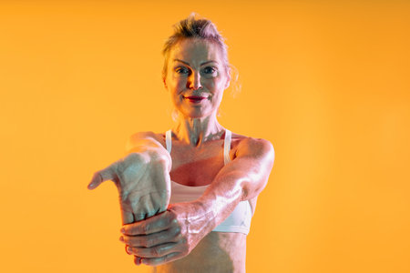 Beautiful mature sportive woman wearing sportswear doing workout - athletic mature woman in sportswear stretching her arms against a vibrant orange backdrop, embodying an active, healthy lifestyleの写真素材