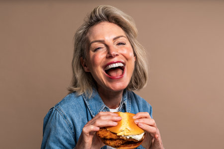 Beautiful mature woman eating tasting hamburger on colorful background - Portrait of pretty middle aged female holding fast food sandwichの写真素材
