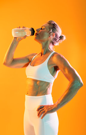 Beautiful mature sportive woman wearing sportswear doing workout - strong mature woman in white sportswear drinking water after an intense workout, embodying a healthy lifestyle for older adultsの写真素材