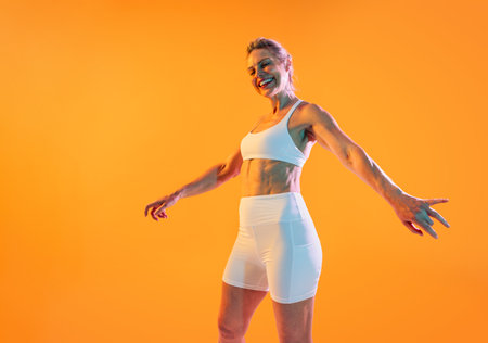 Beautiful mature sporty woman wearing sportswear doing workout - joyfully posing with arms outstretched. To happy. Fit senior woman in white sportswear stands against a vibrant orange backdropの写真素材