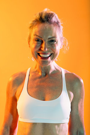 Beautiful mature sportive woman wearing sportswear doing workout - sweaty and smiling mature woman in a white sports bra poses against an orange background, radiating happiness after her workoutの写真素材