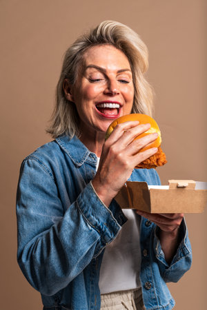 Beautiful mature woman eating tasting hamburger on colorful background - Portrait of pretty middle aged female holding fast food sandwichの写真素材
