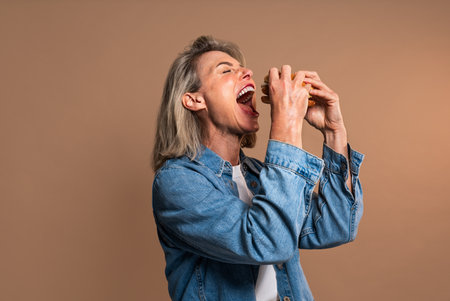 Beautiful mature woman eating tasting hamburger on colorful background - Portrait of pretty middle aged female holding fast food sandwichの写真素材