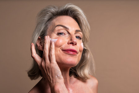 Beauty portrait of beautiful mature woman, concepts about cosmetics, skincare and wellness. Smiling mature woman applying moisturizer to her face. Embracing a skincare routine for healthyの写真素材