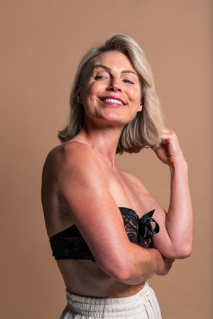 Beauty portrait of beautiful mature woman, concepts about cosmetics, skincare and wellness. Smiling mature woman touching her hair while wearing a black bra and white pantsの写真素材