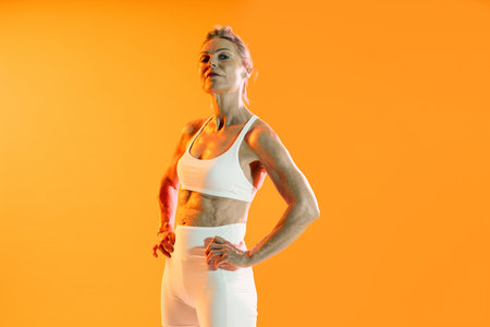 Beautiful mature sportive woman wearing sportswear doing workout - confident senior athlete posing in white sportswear against a vibrant orange background, embodying fitness and active agingの写真素材