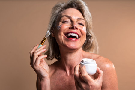 Beauty portrait of beautiful mature woman, concepts about cosmetics, skincare and wellness. Happy. Bare-shouldered mature woman laughing while applying moisturizing face cream with a brushの写真素材