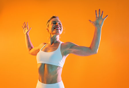 Beautiful mature sportive woman wearing sportswear doing workout - athletic mature woman radiating energy and strength while performing a dynamic workout against a vibrant orange backdropの写真素材