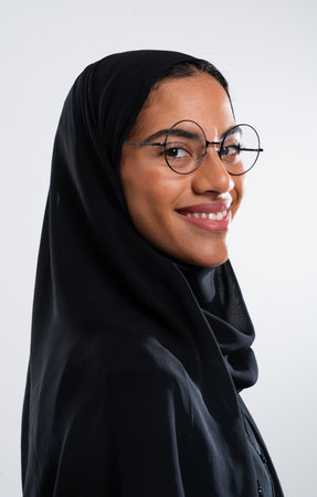 Studio portrait of a smiling arab woman wearing glasses and a traditional black hijab, showcasing...
