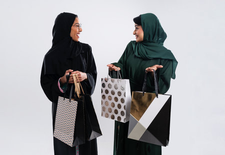 Two smiling arab women wearing traditional abayas are showing shopping bags while talking and having...