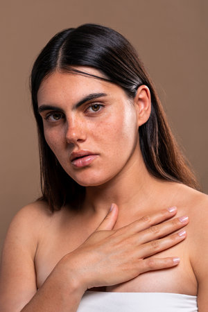 Studio portrait of a young woman with freckles and long brown hair, gently touching her chest,...