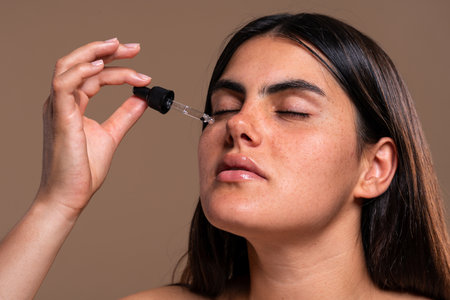 Beautiful young woman with freckles is closing her eyes and applying moisturizing serum on her face...