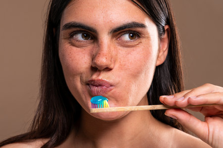 Studio portrait featuring a young woman holding a bamboo toothbrush with blue toothpaste,...