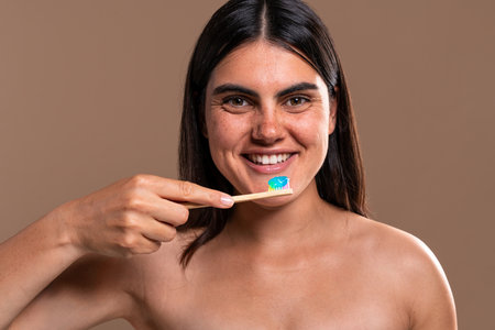 Cheerful young woman with freckles smiles while holding a bamboo toothbrush with rainbow-colored...