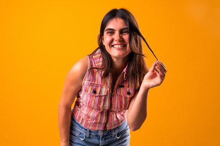 Cheerful young woman playing with her hair while wearing a colorful striped top and denim shorts,...