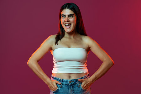 Young woman with tattoo smiling and posing with hands in her pockets, wearing a white top and jeans, in a studio setting with a vibrant, colorful background, expressing confidence and styleの写真素材
