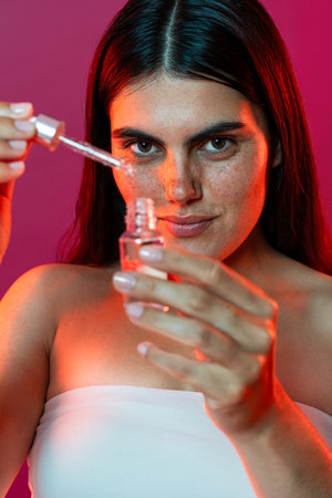 Young woman with freckles holds a glass bottle and dropper, carefully applying face serum as part of her skincare routine in a vibrant studio setting with dynamic lightingの写真素材