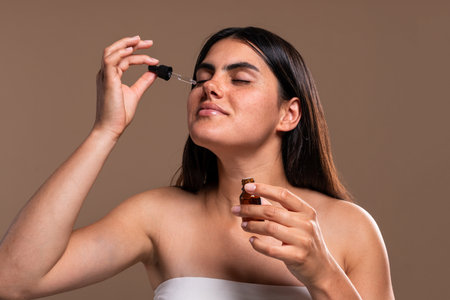 Young woman with closed eyes is applying a face serum using a dropper, holding a small brown bottle in her other hand, demonstrating a skincare routine against a neutral backgroundの写真素材