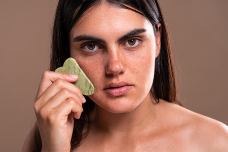 Young woman with freckles gently massages her cheek with a heart-shaped gua sha stone, promoting lymphatic drainage and enhancing her natural beauty as part of a holistic skincare routineの写真素材