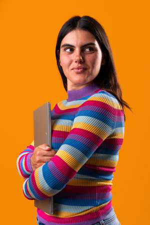 Studio portrait of a young woman with unique style, wearing a colorful striped sweater, holding a laptop and looking away on an orange background, creating a vibrant and modern imageの写真素材