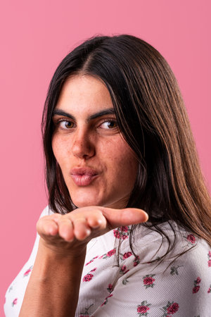 Studio portrait of a young woman with long brown hair and freckles, wearing a white floral shirt, blowing a kiss against a vibrant pink backdrop, expressing affection and playfulnessの写真素材