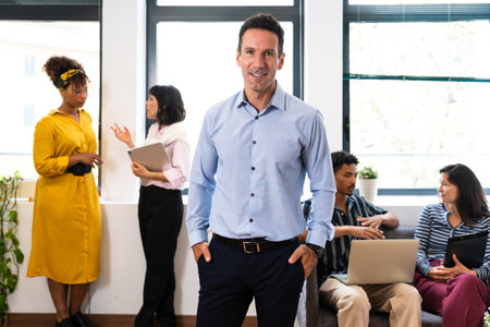 Mature businessman stands confidently with his hands in his pockets, smiling while his diverse team...
