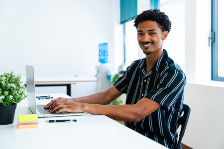 Happy young manager typing on a laptop keyboard while sitting at a desk in a bright modern office,...