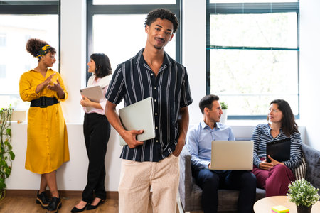 Young, confident businessman holds a laptop while leading a diverse team in a bright, modern startup...