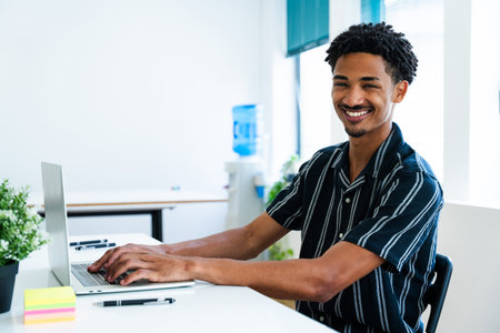 Happy young manager typing on a laptop in a bright startup office, enjoying a modern workplace while...