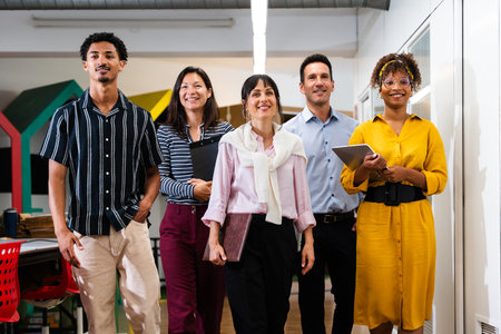 Multi-ethnic business team walking together in a modern office corridor, holding folders and a...