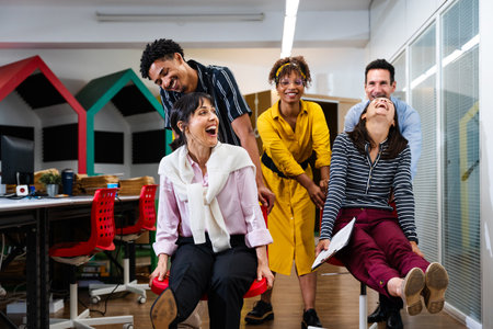 Multi-ethnic businesspeople are enjoying a playful moment, pushing each other on rolling chairs and...