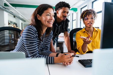 Three smiling business people are collaborating on a project, gathered around a desktop computer in a bright, modern office, discussing strategies and analyzing dataの写真素材