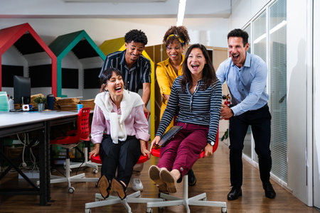 Multi-ethnic business team is enjoying a playful moment, pushing each other on office chairs and...