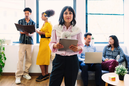 Businesswoman holds a tablet, leading a diverse team collaborating in a bright, modern office,...