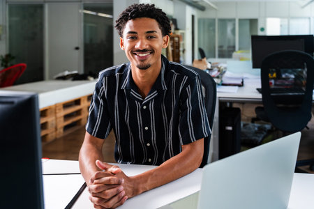 Smiling young manager sitting at his desk in a modern startup office, engaging with both a laptop...