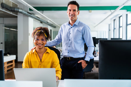 Two smiling colleagues, a woman using laptop and a man with hand in pocket, are working together in...