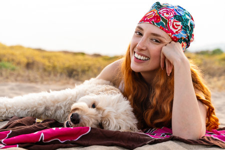 Redhead woman in a colorful bandana lying on a beach towel with her happy doodle dog, enjoying sunny...