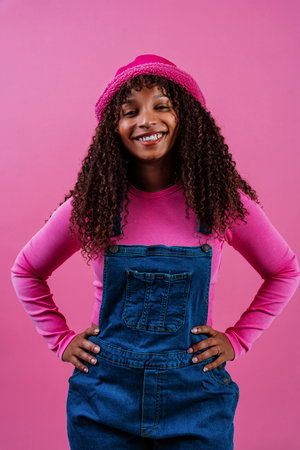 Fashion portrait of a cheerful young woman with curly hair, wearing a pink bucket hat, pink long-sleeved shirt and denim overalls, laughing over a pink backgroundの写真素材