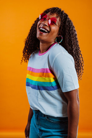 Studio shot of a cheerful young woman wearing rainbow t-shirt and red sunglasses showing peace sign...