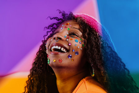 Studio portrait of a cheerful young woman with curly hair, wearing colorful rhinestones on her face,...