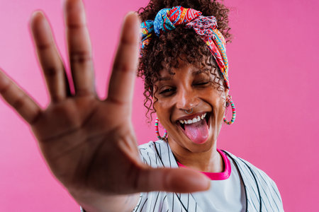 Studio shot of a cheerful young woman with colorful headscarf and rainbow striped shirt showing her open hand and smiling against a vibrant pink backgroundの写真素材