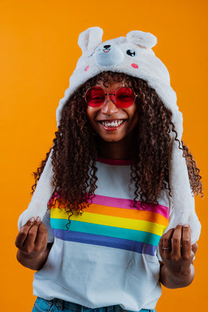 Young woman with curly hair wearing a funny hat and rainbow t-shirt is laughing and having fun on an orange background, celebrating diversity and inclusionの写真素材
