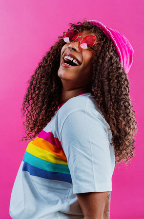 Studio shot of a cheerful young woman with long curly hair wearing a rainbow t-shirt, yellow...