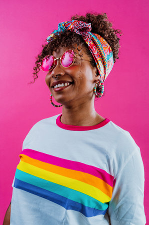 Cheerful young woman with curly hair, wearing a colorful headscarf and pink sunglasses, smiling and...