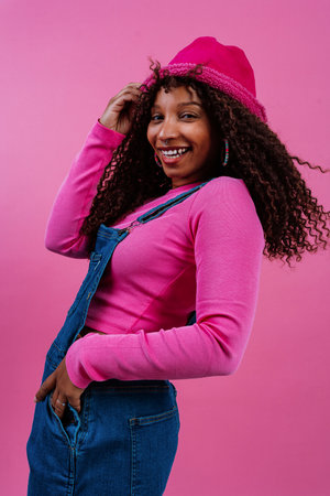 Fashion portrait of a cheerful young woman with curly hair, wearing a pink bucket hat, pink long-sleeved shirt and jeans, laughing with colorful rhinestones on her face over a pink backgroundの写真素材