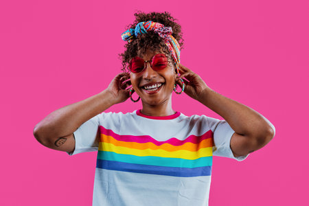 Cheerful young woman with curly hair, wearing a colorful headscarf and pink sunglasses, smiling and looking away against a vibrant pink background, celebrating diversityの写真素材