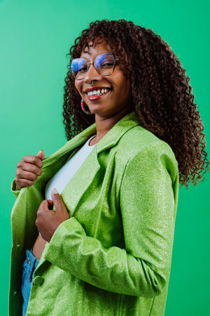 Studio portrait of a cheerful young woman with curly hair laughing wearing eyeglasses and green jacket, standing with hand in pocket on a green backgroundの写真素材