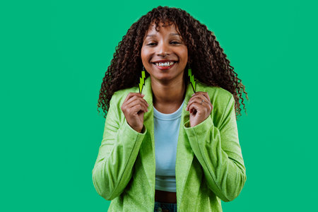 Studio portrait of a cheerful young woman with curly hair, wearing a green jacket, showing her green lightning bolt earrings and smiling on a green backgroundの写真素材