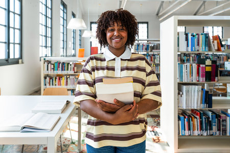 Young black student standing and smiling in a modern library, holding a stack of textbooks while...